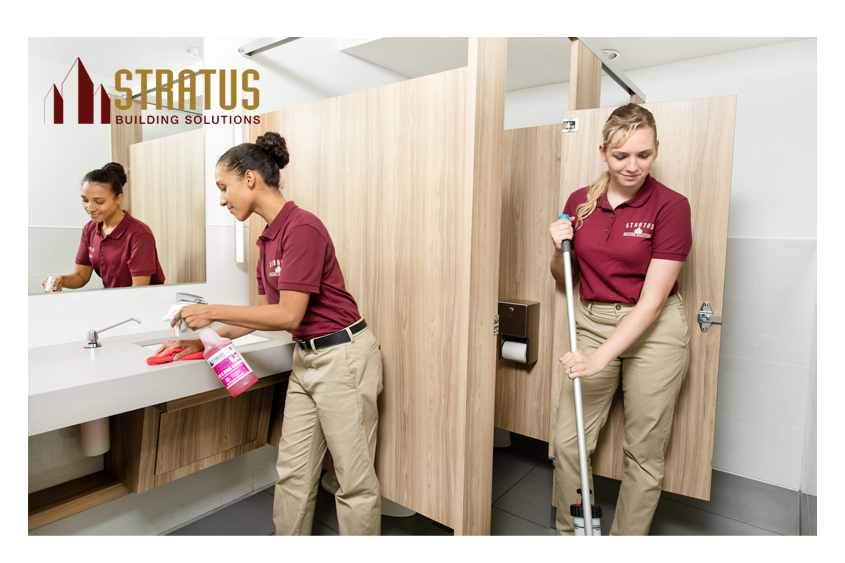 A Uniformed Stratus Employee Sprays and Wipes Down a Bathroom Counter While Another Exits a Stall While Mopping the Floor